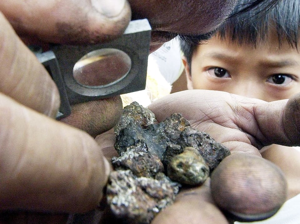 Ein Mann schaut mit einer Lupe einen verdreckten Klumpen nach Gold ab. Im Hintergrund schaut ein Kind auf das Gestein.