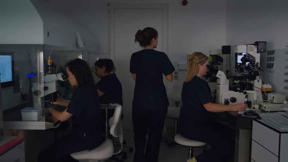 Ein dunkles Labor, drei Frauen sitzen konzentriert an den Mikroskopen, eine Frau ist von hinten zu sehen und läuft aus dem Bild.