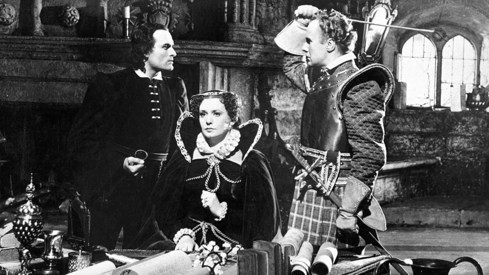 Eine Szene aus einem alten Film: Eine Königin in prunkvollem Gewand sitzt an einem Tisch, links und rechts von ihr stehen zwei Männer und schauen sich in die Augen. Die Königin schaut starr geradeaus.