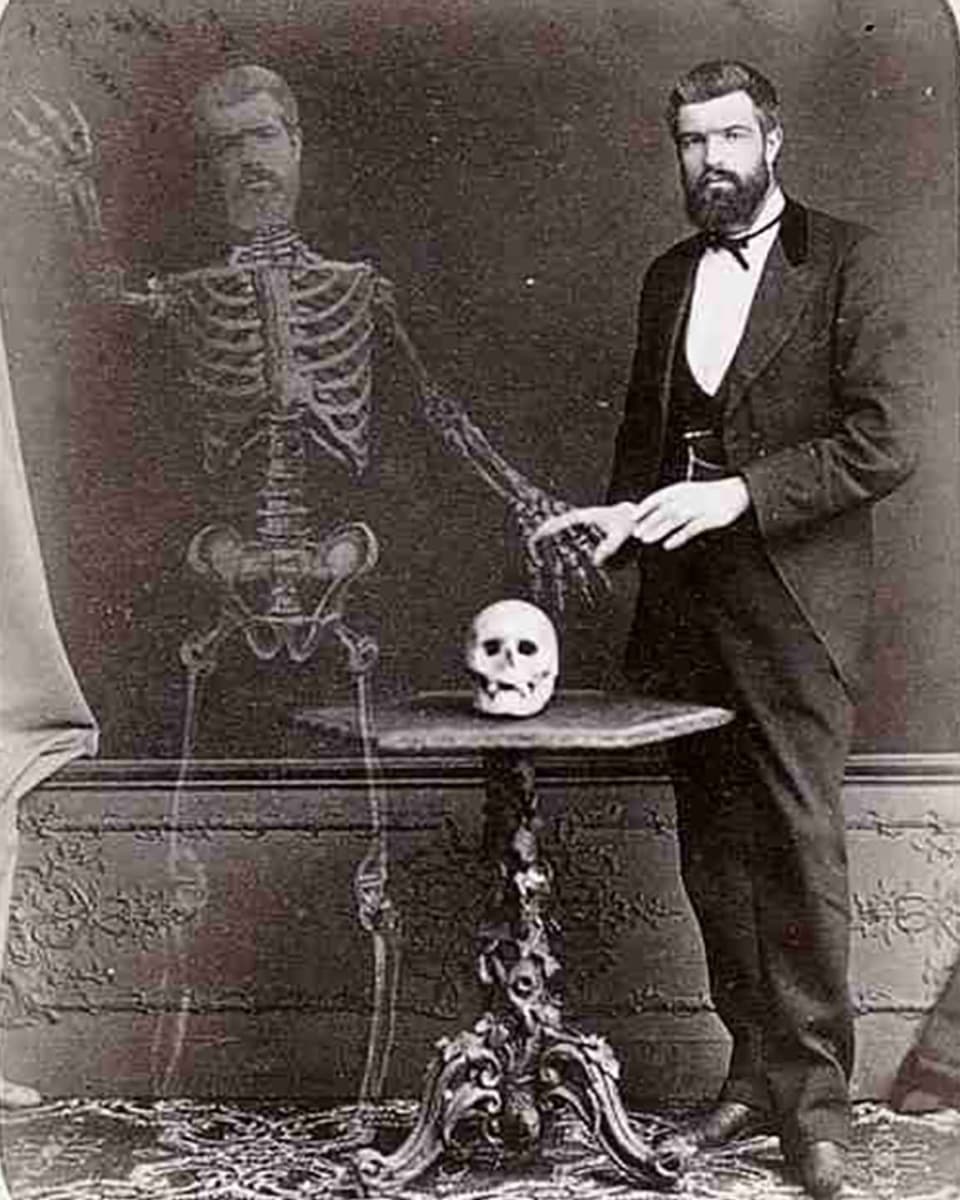 Schwarzweissfoto: Ein viktoranischer Mann steht neben einem Skelett, dessen Kopf sein eigener ist.