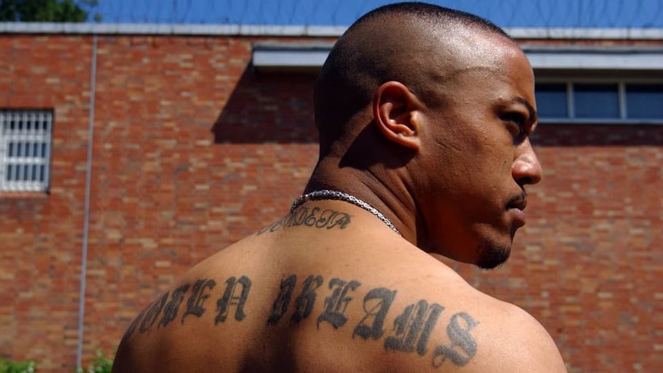 Mann mit Glatze, Kette und zahlreichen Tattoos in Rückansicht vor Gefängnismauer