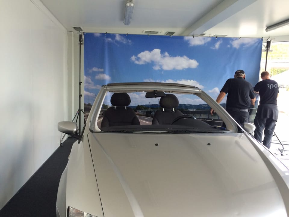 Ein Auto von vorne, dahinter wird eine Leinwand montiert, die einen Himmel zeigt.