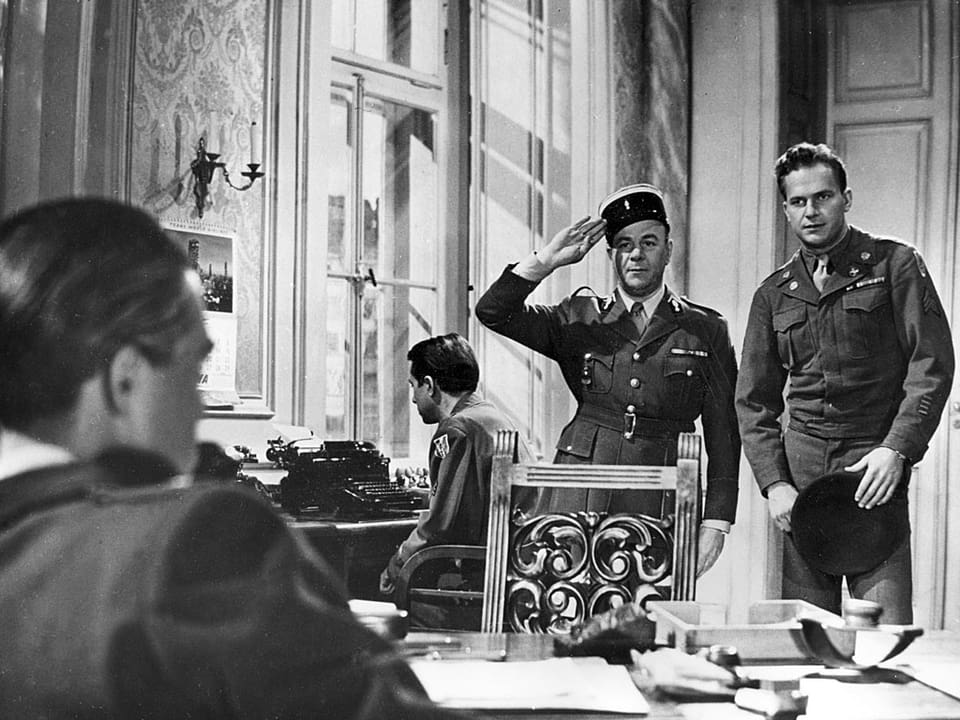 Zwei Soldaten stehen vor einem Mann, der an einem Tisch sitzt.