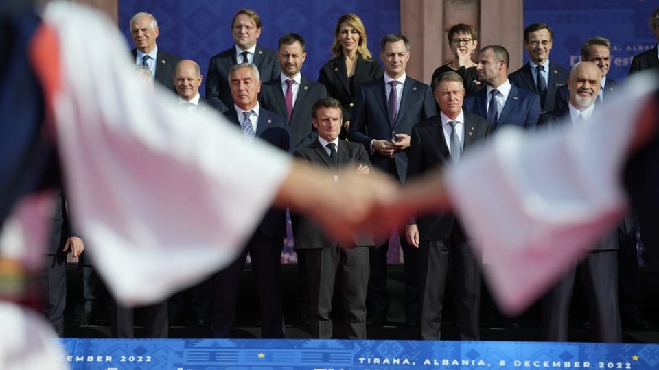Zwei Personen geben sich im Vordergrund die Hände und im Hintergrund stehen die EU-Staats- und Regierungschefs