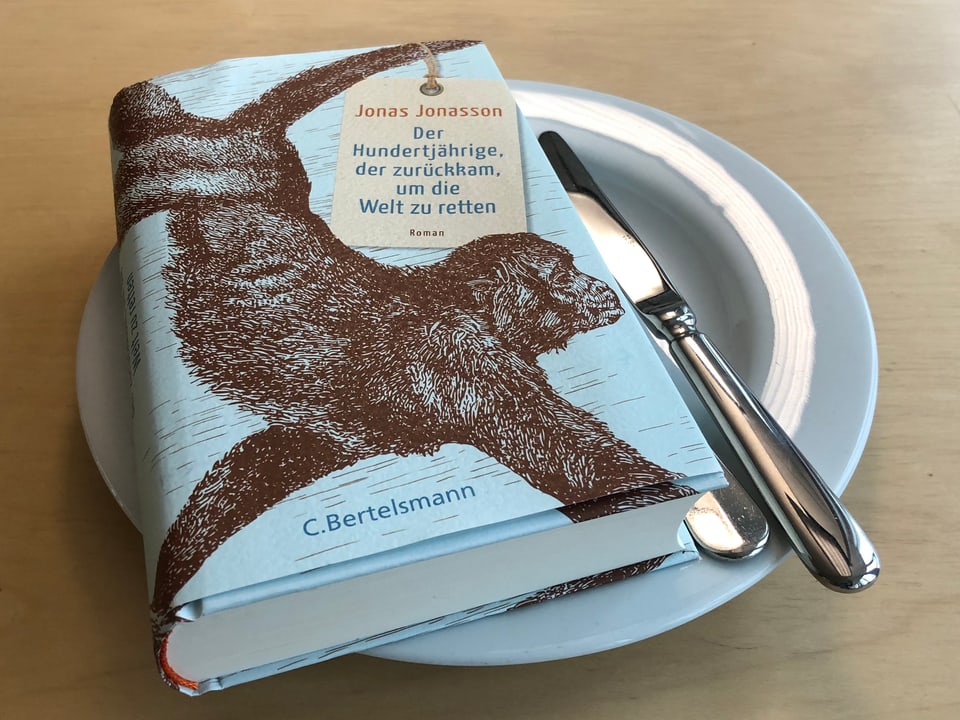«Der Hundertjährige, der zurückkam, um die Welt zu retten» liegt auf einem weissen Teller. Messer und Gabel seitlich neben das Buch auf den Teller gelegt.