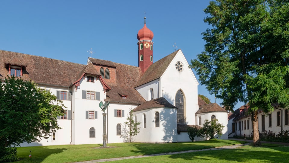 Ein Kloster mit Turm und Wiese und Laubbaum bei schönem Wetter.
