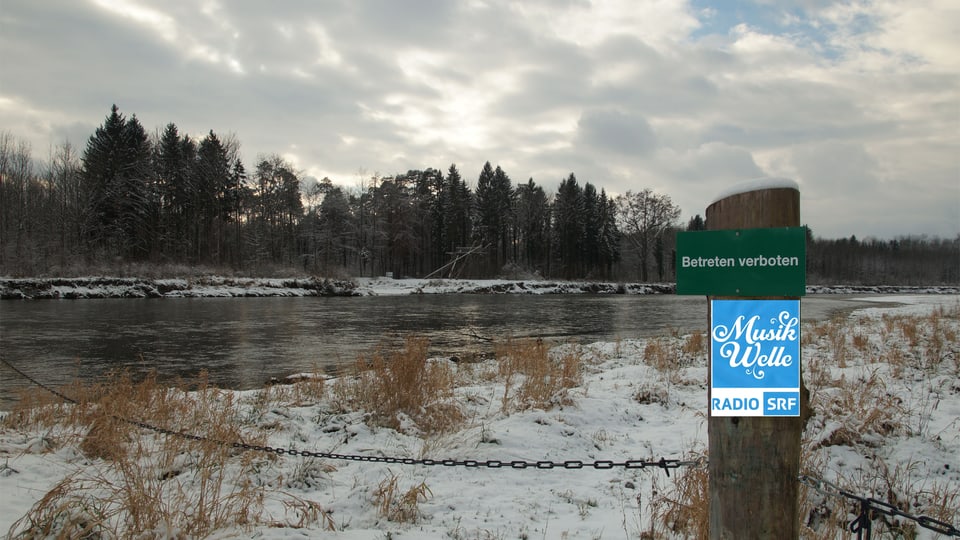 Das Logo von SRF Musikwelle an einem Pfosten, der in einer kalten Winterlandschaft an einem Fluss steht.