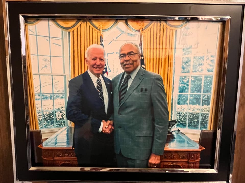 Foto eines gerahmten Fotos, darauf links Joe Biden und rechts älterer, schwarzer Mann im Anzug, dahinter das Oval Office