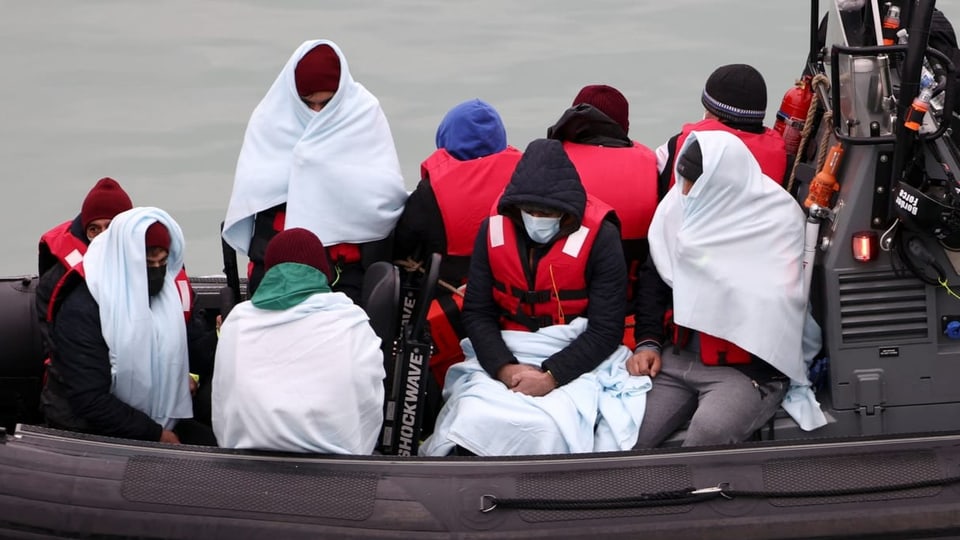 Ungefähr neun männliche Flüchtlinge sitzen in einem Boot, eingehüllt mit weissen Decken.