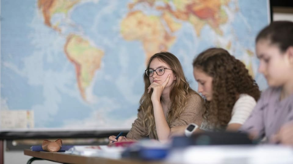 Drei junge Frauen sitzen bei einer Prüfung vor einer Weltkarte. 