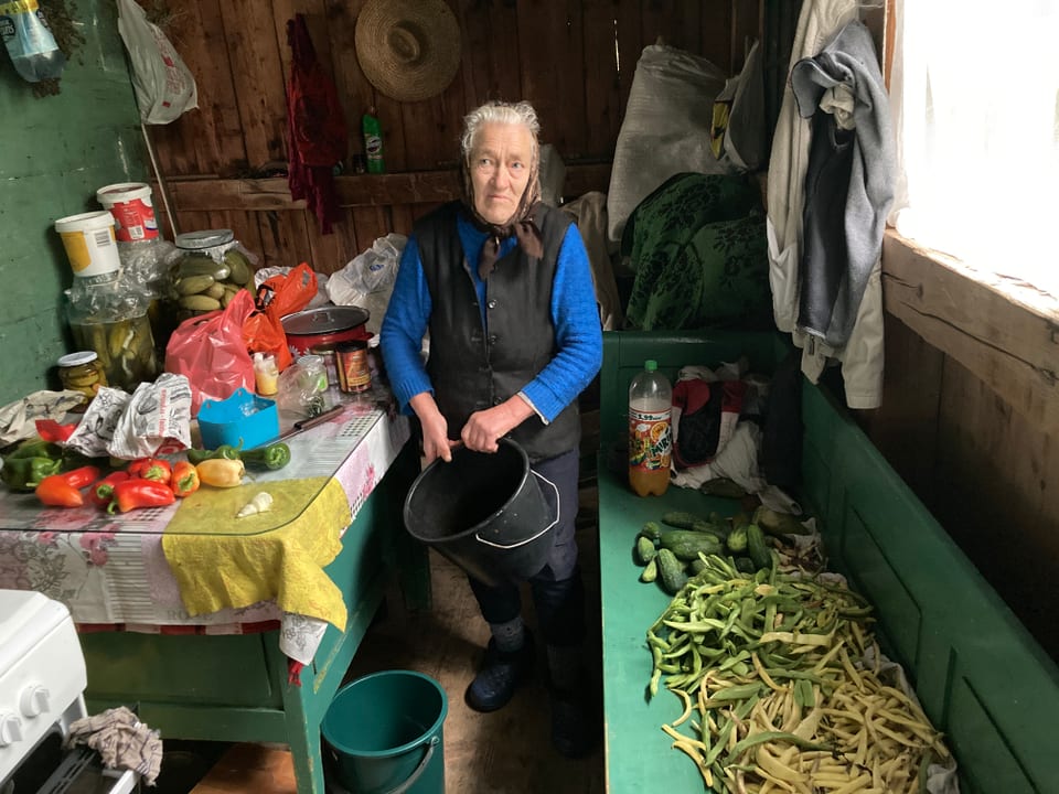 Ältere Frau in Kopftuch hält einen schwarzen Topf. Sie steht in ihrer Küche. Auf der Seite sieht man Gemüse. 