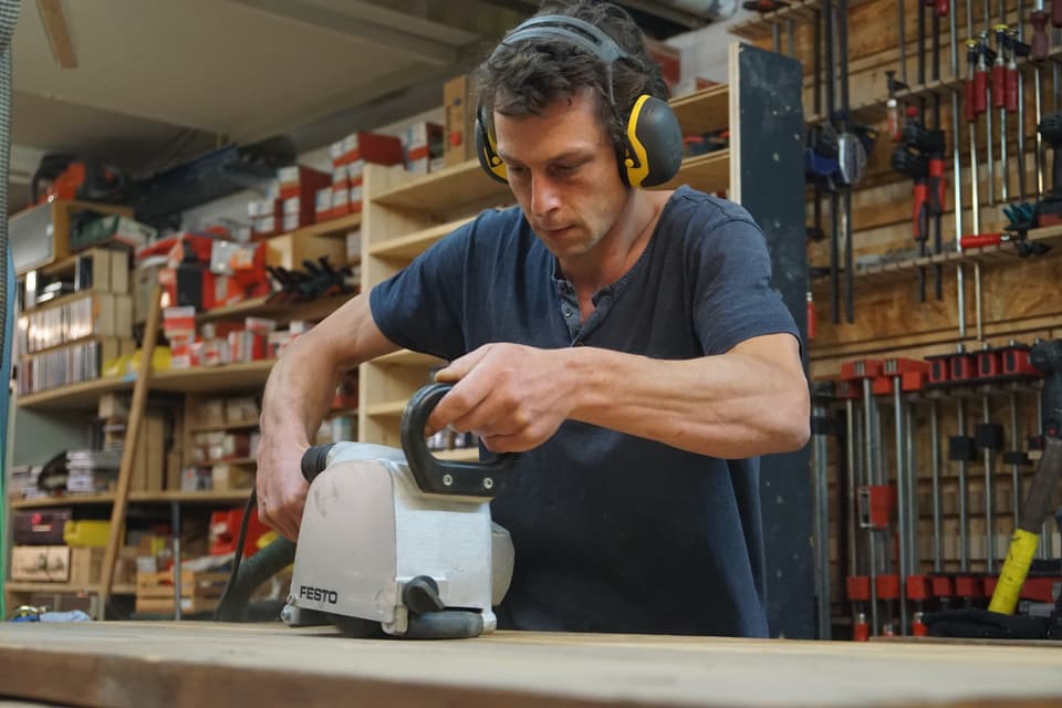 Ein Mann mit Gehörschutz bearbeitet eine Holzplatte mit einer Schleifmaschine.
