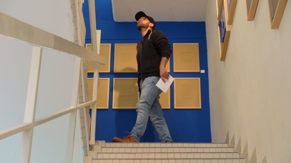 Ein junger Mann mit Jeans, dunklem Pullover und einem Cap auf dem Kopf in einem Treppenhaus.