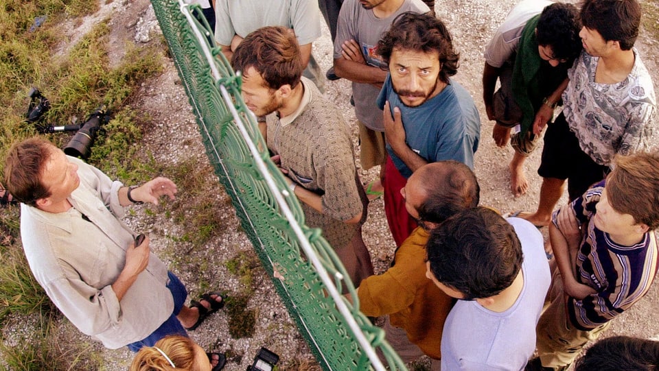 Flüchtlinge auf Nauru von oben fotografiert diskutieren mit einem Mann durch einen Zaun