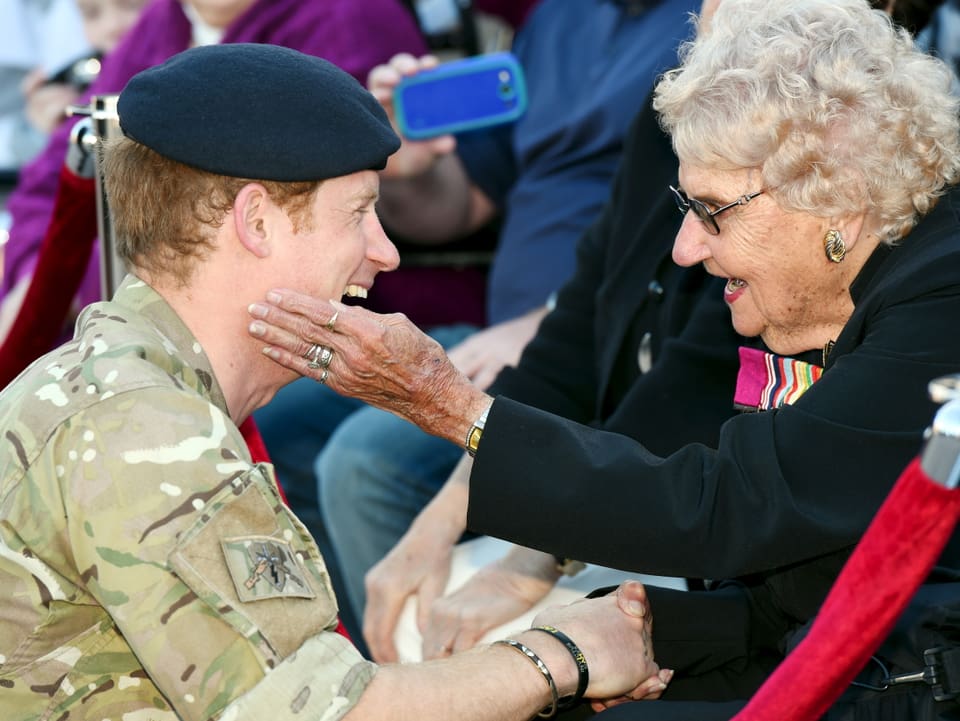 Eine ältere Dame streichelt Prinz Harry mit der Hand über seine Wange.