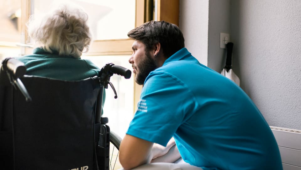 Junger Mann spricht mit alter Frau in einem Rollstuhl.