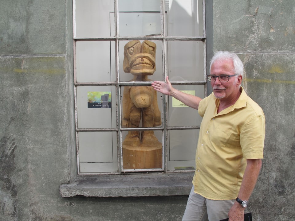 Hans Estermann steht vor einem Fenster hinter dem eine Holzskulpur ausgestellt ist.