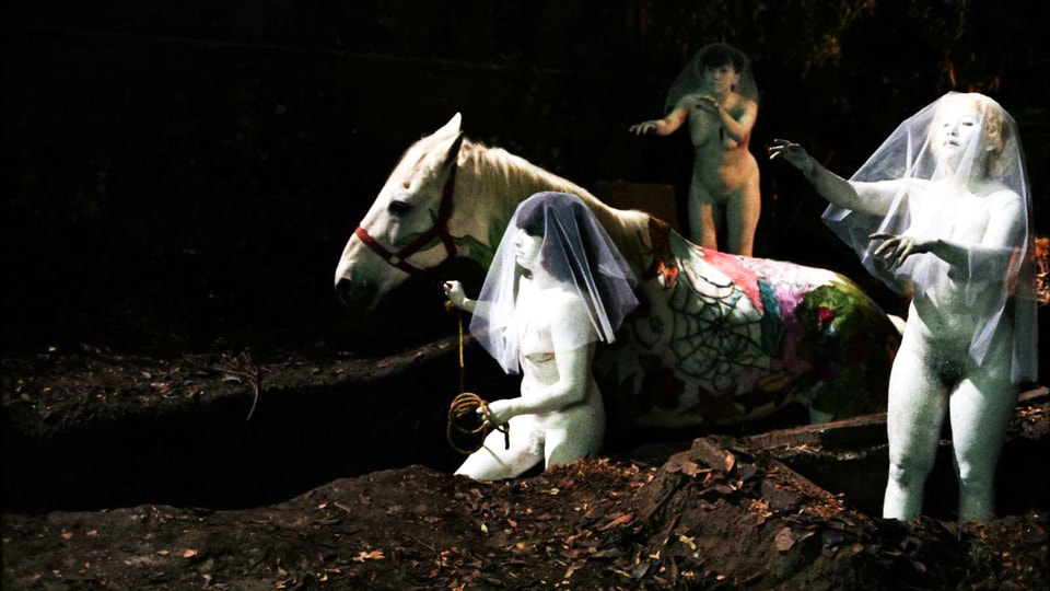 Nackte Frauen mit Brautschleier führen ein Pferd durch die Dunkelheit.
