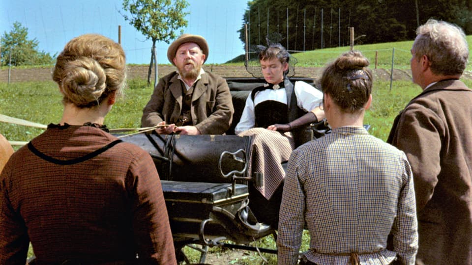 Ein Mann mit Vollbart sitzt auf einer Kutsche. Neben ihm sitzt eine junge Frau mit Schleier. Im Vordergrund stehen zwei Frauen und ein Mann, welche zur Kutsche blicken.