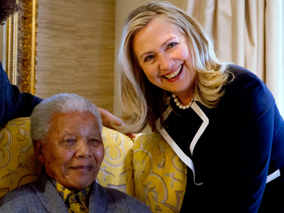 Hillary Clinton beugt sich für ein Foto zu Mandela, der sitzt. Beide lächeln.
