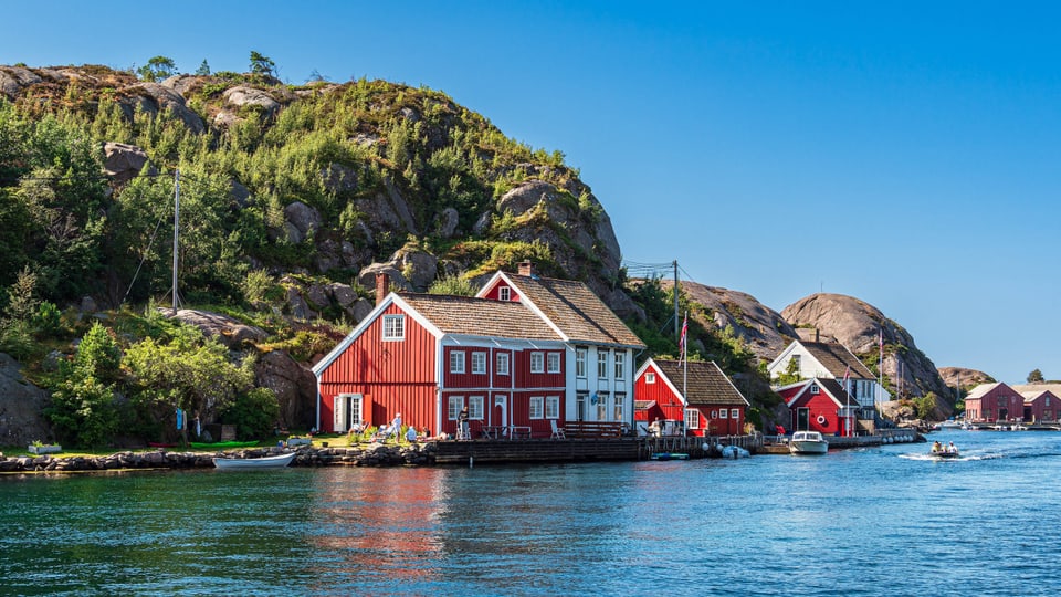 Rot-weisse Häuser in Norwegen, direkt am Wasser. Dahinter Büsche und Felsen.