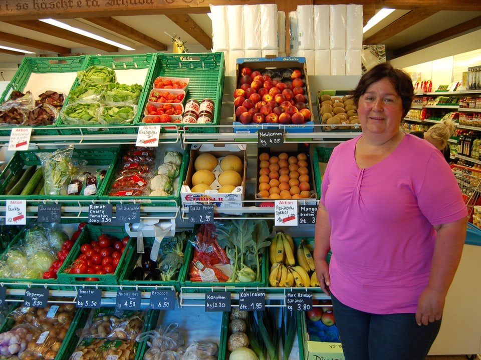 Die Gschäftsführerin trägt ein rosa T-Shirt und Jeans und steht vor dem Früchte- und Gemüseregal.