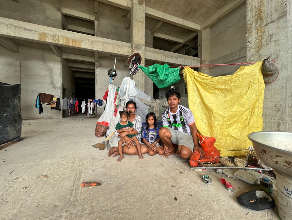 A woman and a man sit on the ground inside a building envelope with their two children.  They hung tarps.