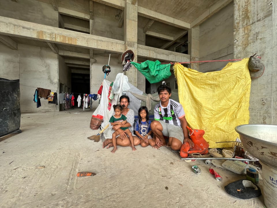 A woman and a man sit on the ground inside a building envelope with their two children. They hung tarps.