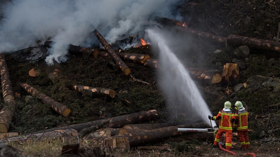Zwei Personen der Feuerwehr spritzen Wasser auf brennendes Holz.