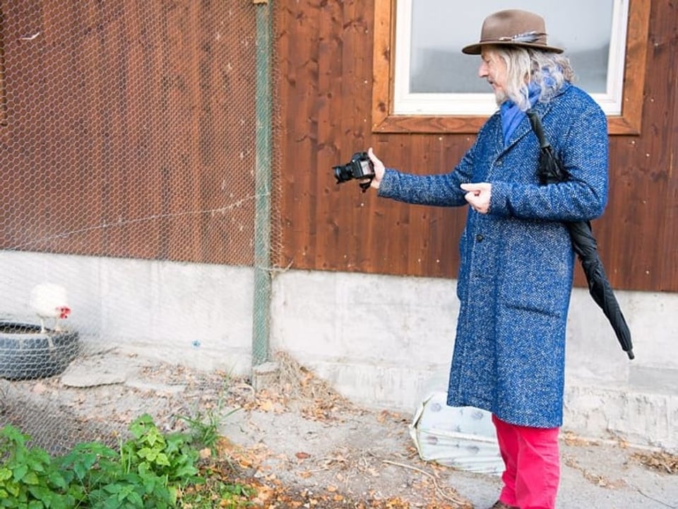 Ein älterer Mann in blauem Mantel und braunem Hut, einen Hut auf dem Kopf und einem Schirm unter dem Arm beim Fotografieren.