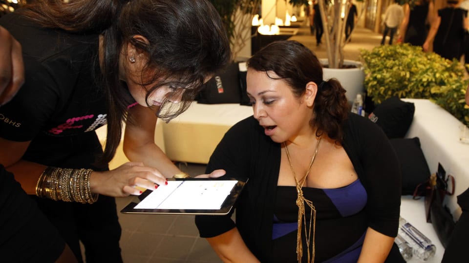 Eine Frau mit einem Tablet-Computer in der Hand beugt sich zu einer ihr rechts gegenübersitzenden Frau hinunter.