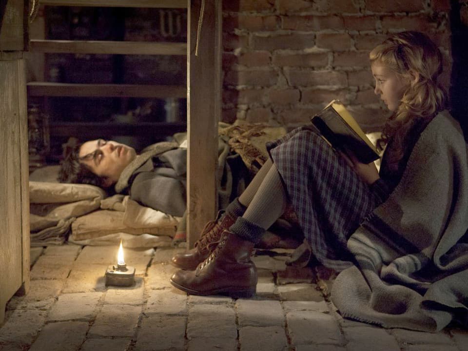 Mädchen sitzt auf Kellerfussboden und liest krankem Mann, der ebenfalls auf dem Boden liegt, aus einem Buch vor.