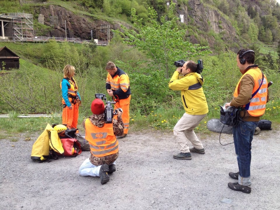 Zwei Kameraleute, ein Tonoperateur, eine Moderatorin und ein Bergführer stehen mit gelben und orangen Warnkleidern auf einem Platz. Die Kameraleute sind am Filmen.