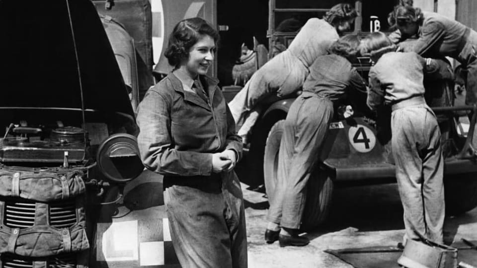 Eine junge Frau steht vor einem Panzer, im Hintergrund reparieren vier Leute ein anderes Fahrzeug.