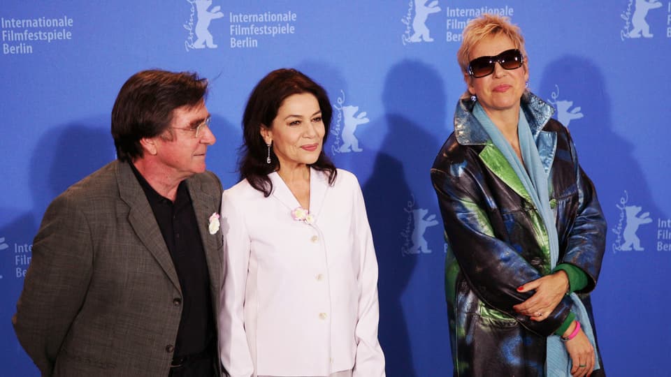 Ein älteter Mann und zwei Frauen stehen vor einer Wand mit dem Berlinale-Logo.