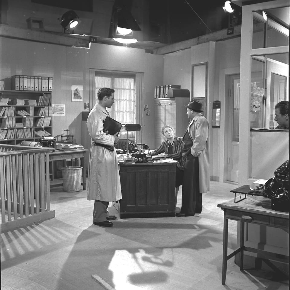Zwei männliche Schauspieler stehen an einem Schreibtisch an dem eine Schauspielerin sitz. Oberhalb der Kulisse sind Filmscheinwerfer angebracht.