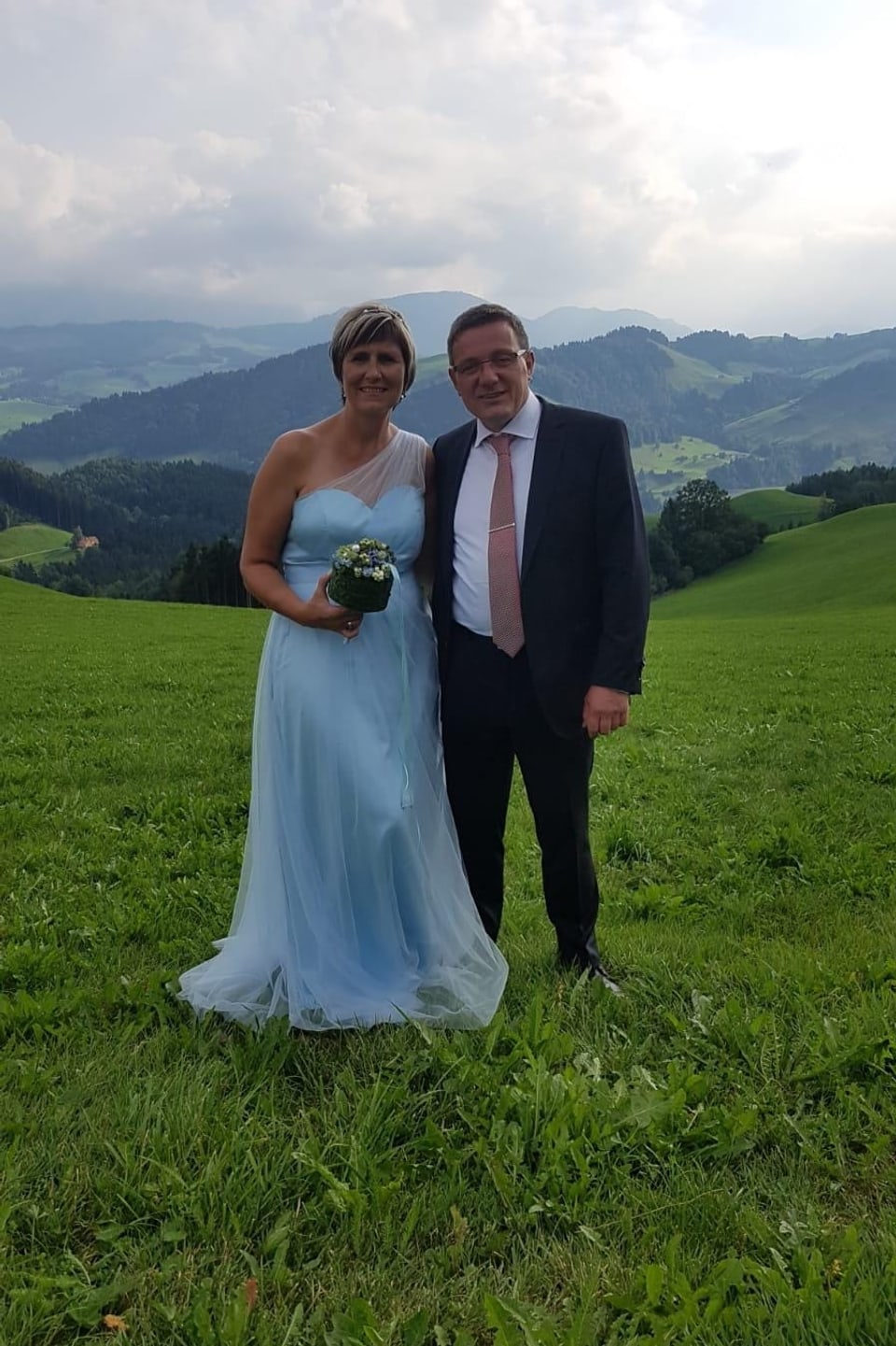 Ein Brautpaar, sie in hellblauem Kleid, er mit Anzug und Kravatte, stet auf einem grünen Feld, im Hintergrund hügelige Landschaft.