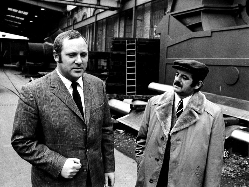 ein Mann mit Kravatte und Anzug und ein Mann mit Beret und Mantel stehen am Bahnhof
