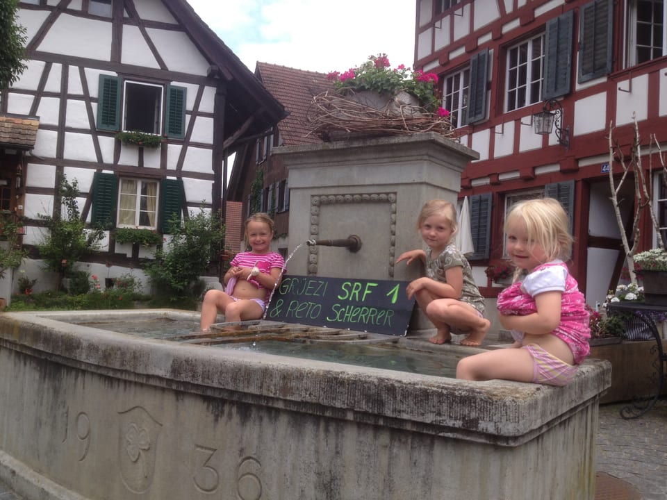 Die Mädchen sitzen auf dem Brunnenrand, ihre Beine im Brunnen. Am Brunnen lehnt ein Schild mit Willkommensgrüssen.