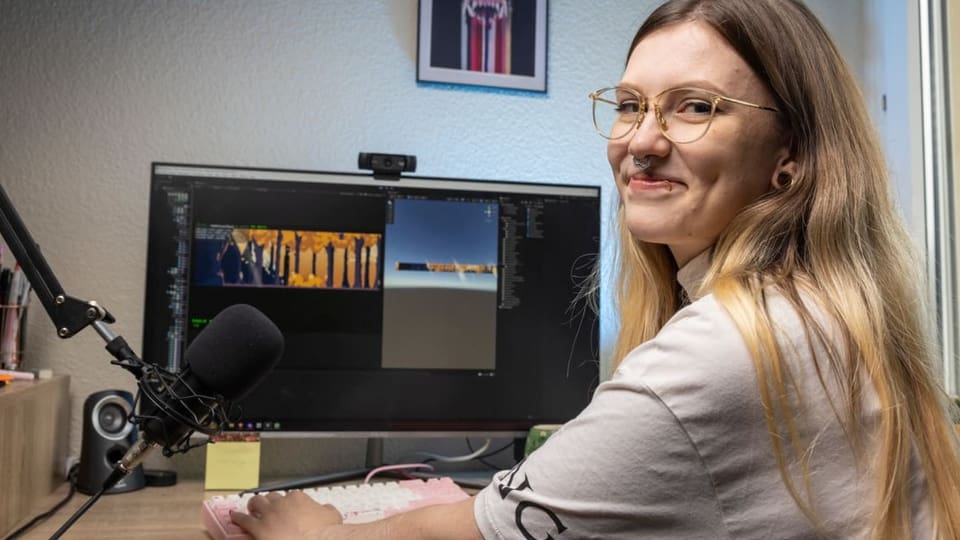 Junge Frau mit langem, blondem Haar, Brille, Nasenring und Lippenpiercing am PC, dreht sich zu Kamera hin, lächelt.