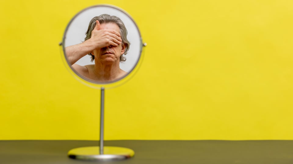 Ein Mann mit welligen Haaren schaut in einen runden Spiegel. Er hält sich die Hand vor das Gesicht