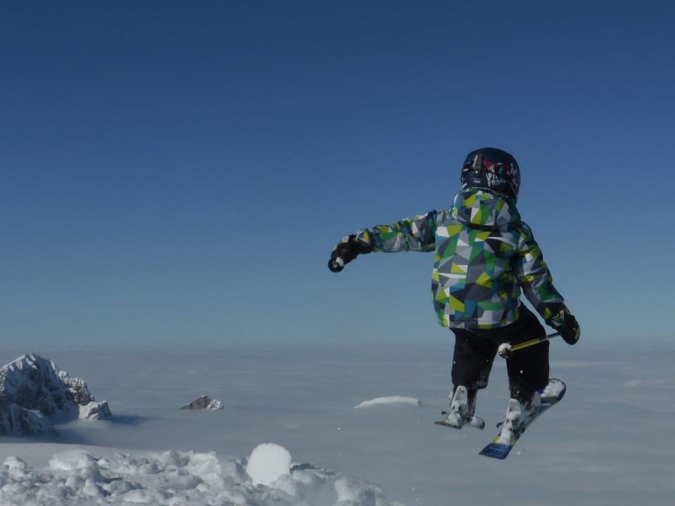 Junge in Skimontur springt mit Skier in die Luft. Er scheint vom sonnigen Berg hinunter auf das Nebelmeer zu springen. 