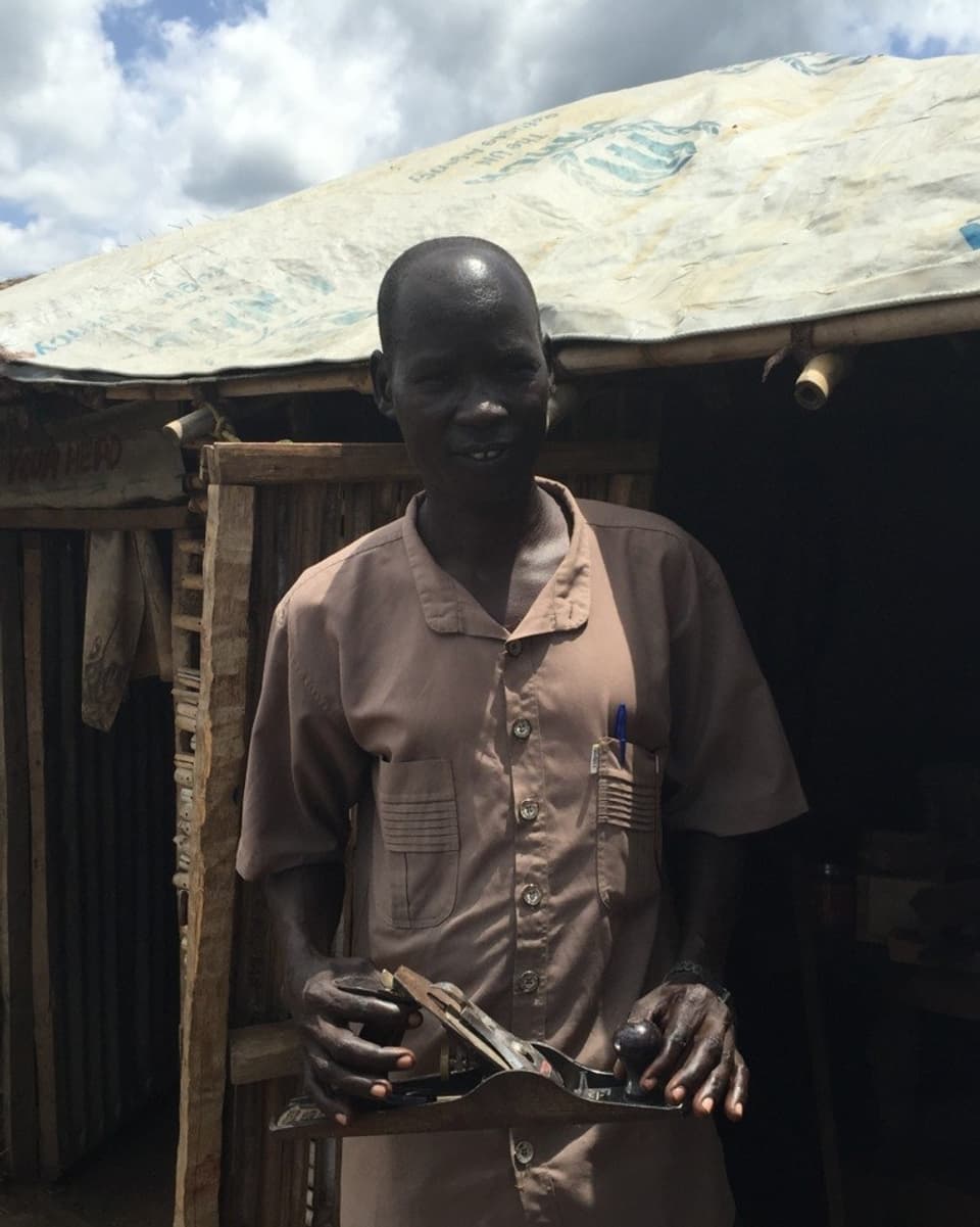 Abuyi Hassan steht vor seinem Häuschen mit einem Hobel in der Hand.