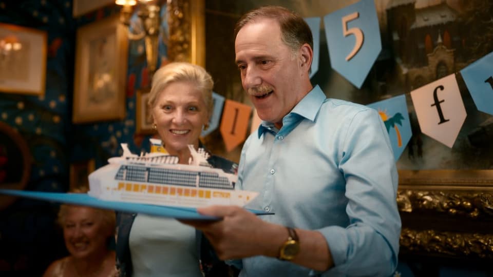 zwei ältere Menschen, links eine Frau, rechts ein Mann, schauen mit überraschtem Ausdruck auf Karton-Schiff