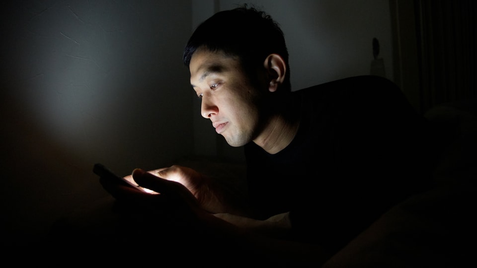 Ein asiatischer Mann in einem dunklen Raum starrt auf sein Handy.