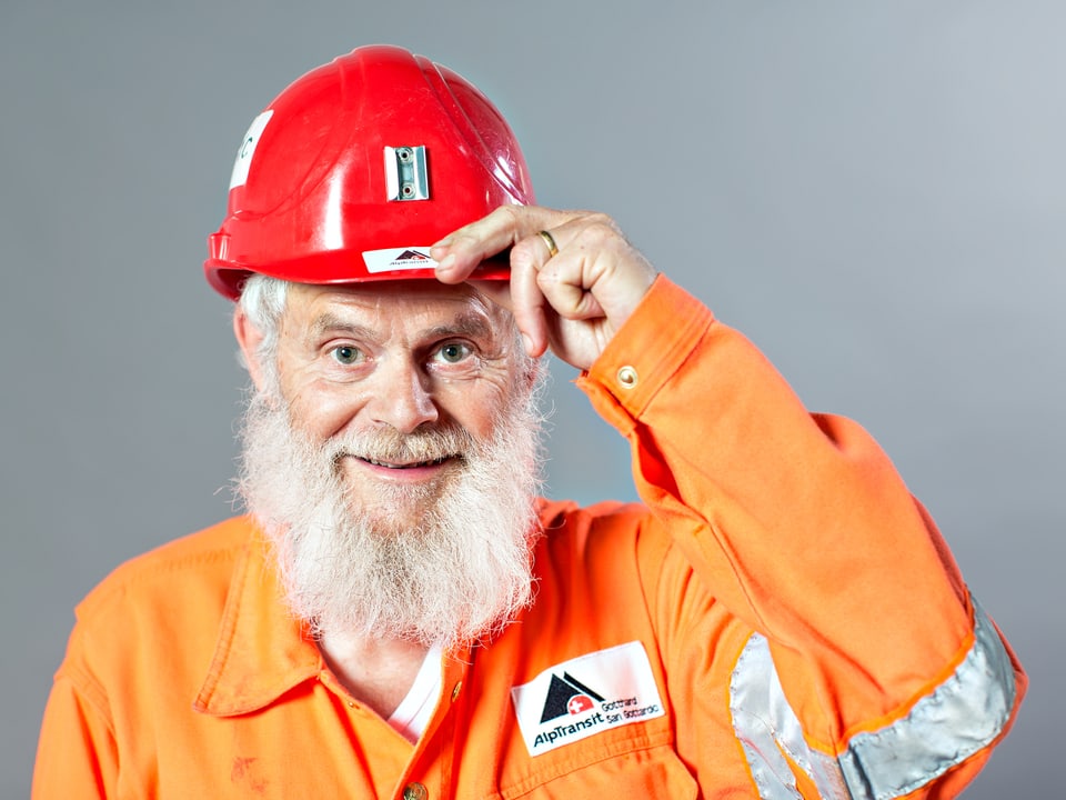 Älterer Mann mit weissem Bart, der seine Hand an den roten Helm auf seinem Kopf hält.