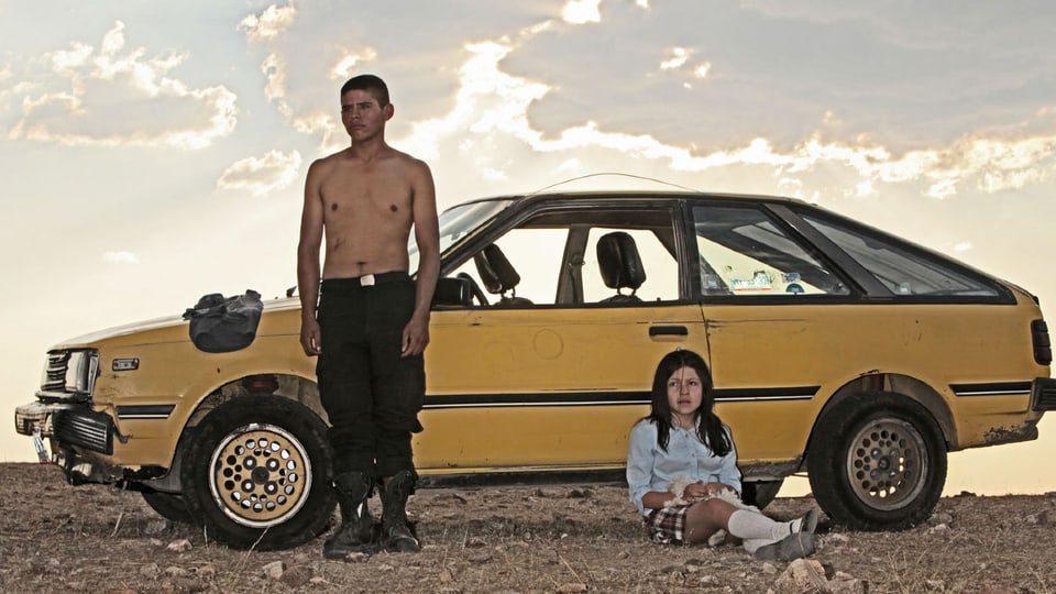 Der Mexikaner Heli, die Hauptfigur in Amat Escalantes Film angelehnt an ein Auto in der mexikanischen Wüste, danben sitzt ein Mädchen.
