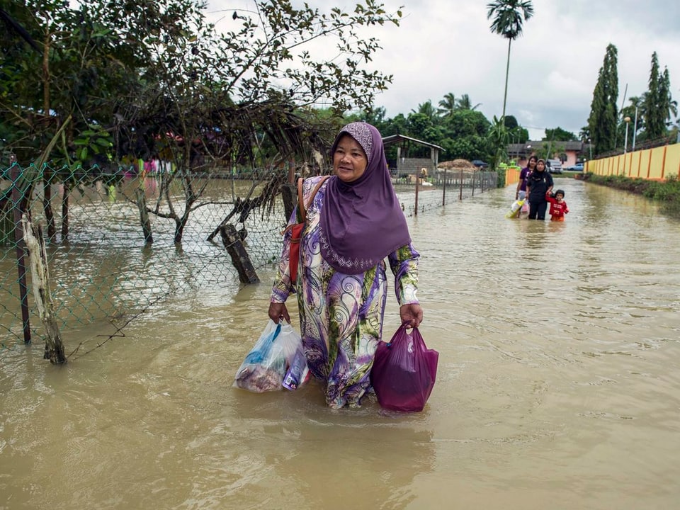 Eine Frau mit zwei Taschen in der Hand geht auf einer überfluteten Strasse. 