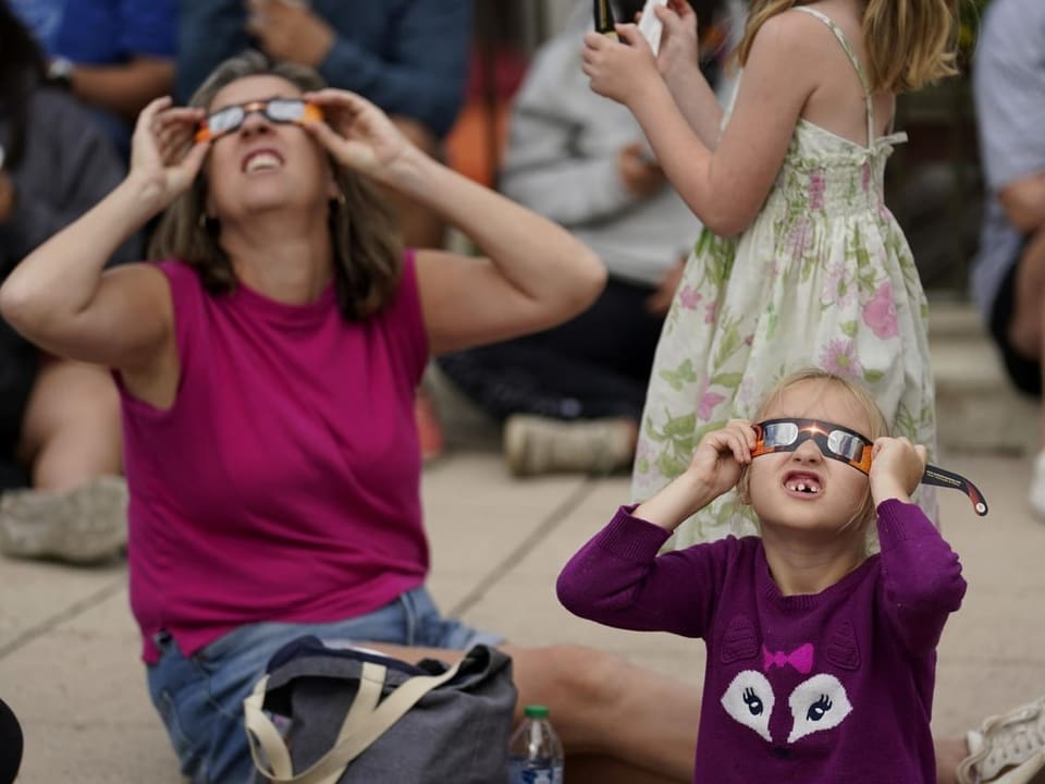 Eine Frau und ein Kind sitzen nebeneinander auf dem Boden. Sie tragen Schutzbrillen und schauen in den Himmel.