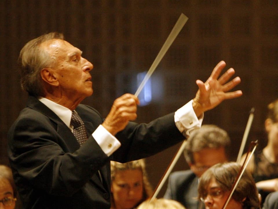 Ein älterer Mann mit lichtem Haar und in schwarzem Anzug hält in der rechten Hand einen Dirigentenstab.